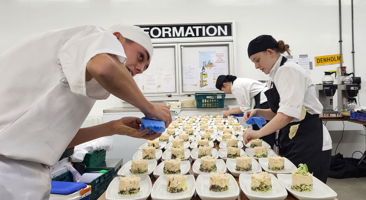 Three Culinary Arts students in chef uniforms put the finishing touches to a table full of plated dishes 